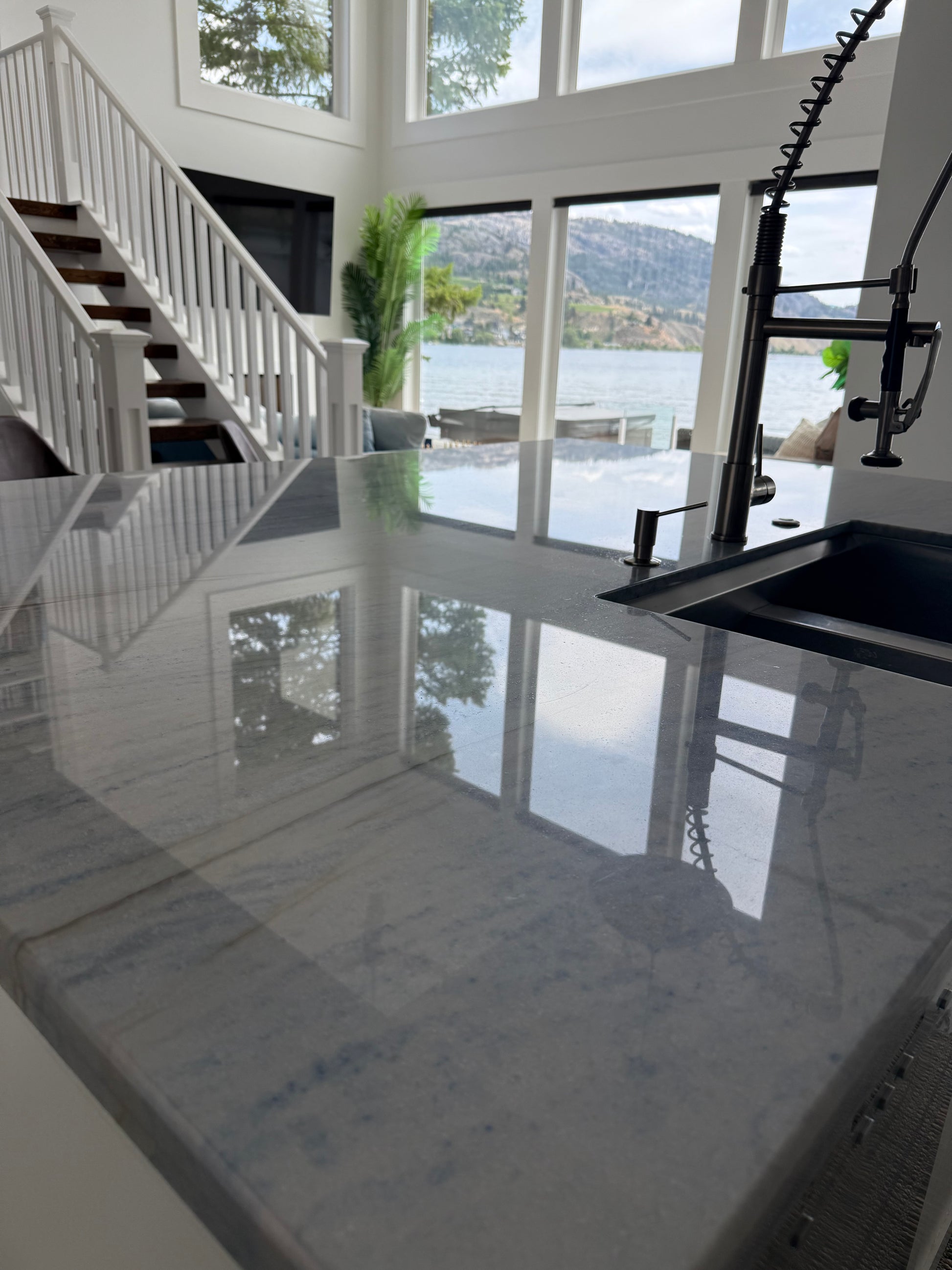 A close up of a kitchen counter that has just been cleaned so well you can see the refelction of the staircase to the left and the lake and hills through the windows that are seen in the distance face on. To the right of the picture we see a blank sink with black tap fixtures.