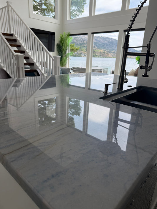 A close up of a kitchen counter that has just been cleaned so well you can see the refelction of the staircase to the left and the lake and hills through the windows that are seen in the distance face on. To the right of the picture we see a blank sink with black tap fixtures.
