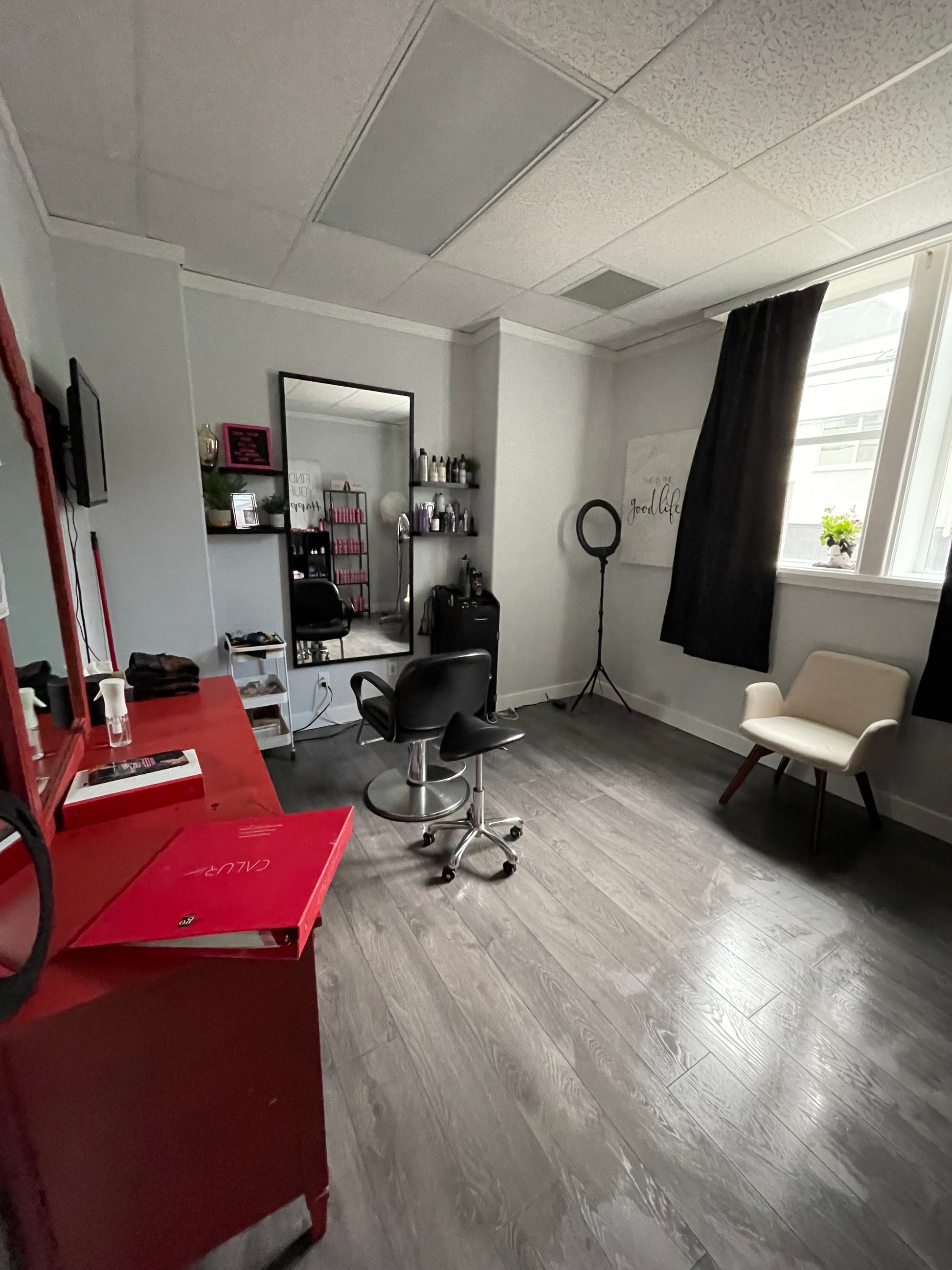 In a room with shining laminate floors after being expertly cleaned, there is a red dresser and mirror which can be seen to the left from the side on one wall, and on the wall next to that is a large mirror with a hairdresser chair set up in front of it. There is a ring light in one corner, and the final wall has a window with a curtain and a small white upholstered chair underneath.