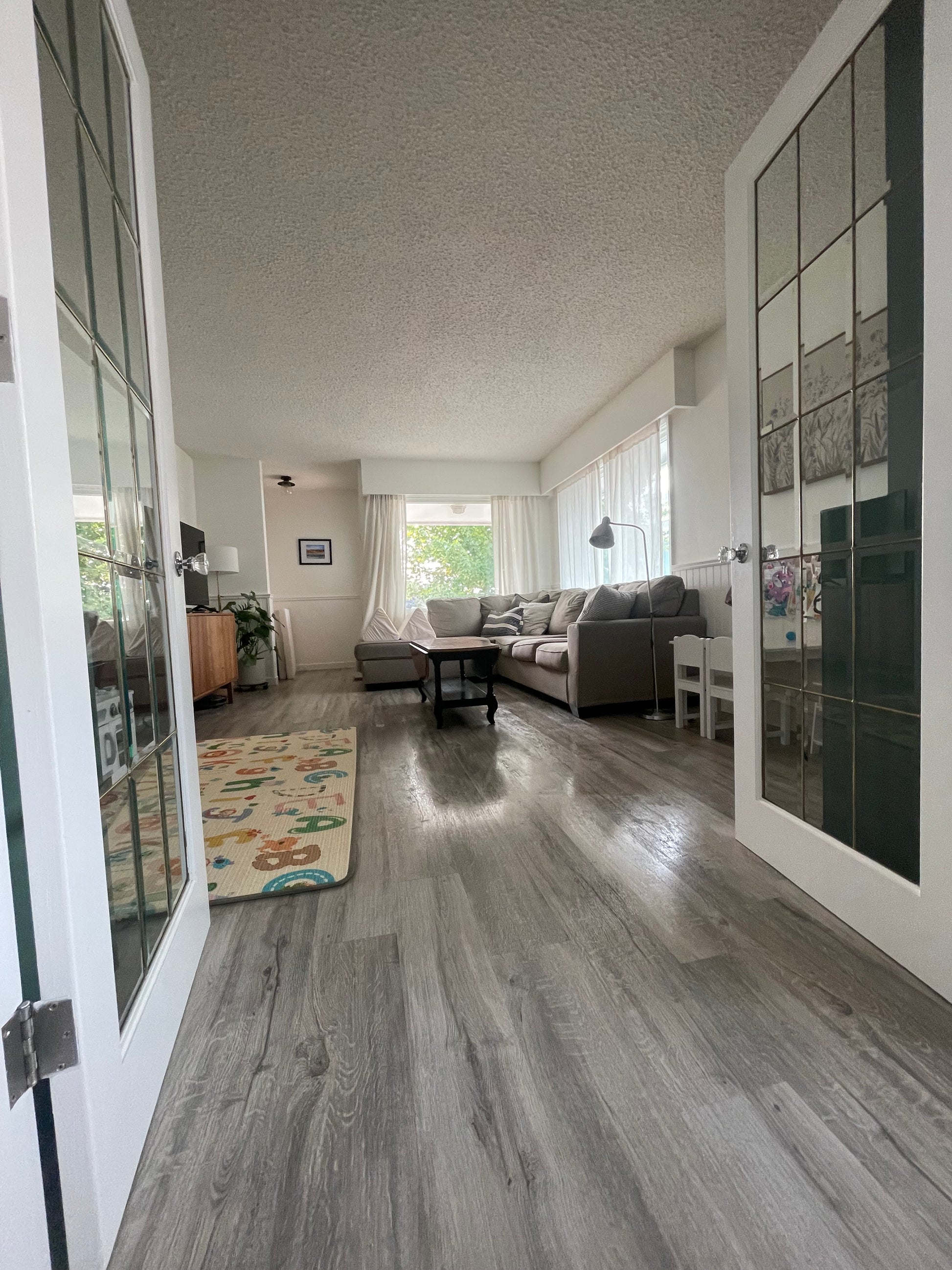 The picture is viewed through open french doors with a shining hardwood floor after it has just been cleaned looking into a living room with a couch, coffee table and windows.