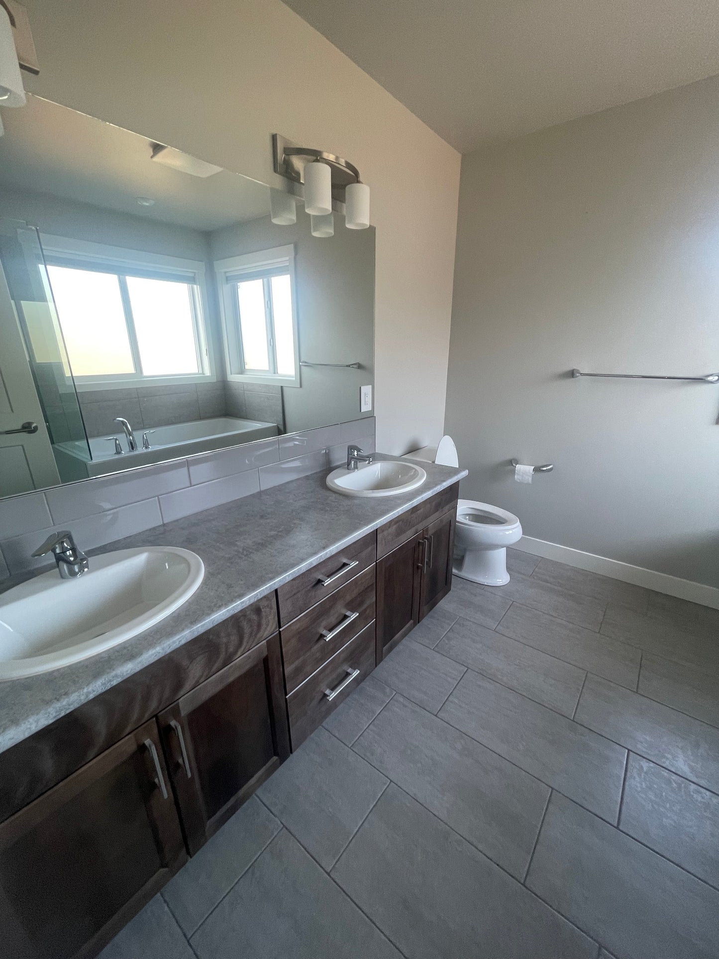 This picture appears to be of a bathroom with a double sink vanity that has grey countertops, dark grey cabinets, a large mirror that is so clean you can see the reflection of a white bathub and light coming in through the windows. We see a toilet, toilet paper and a towel bar in the distance.