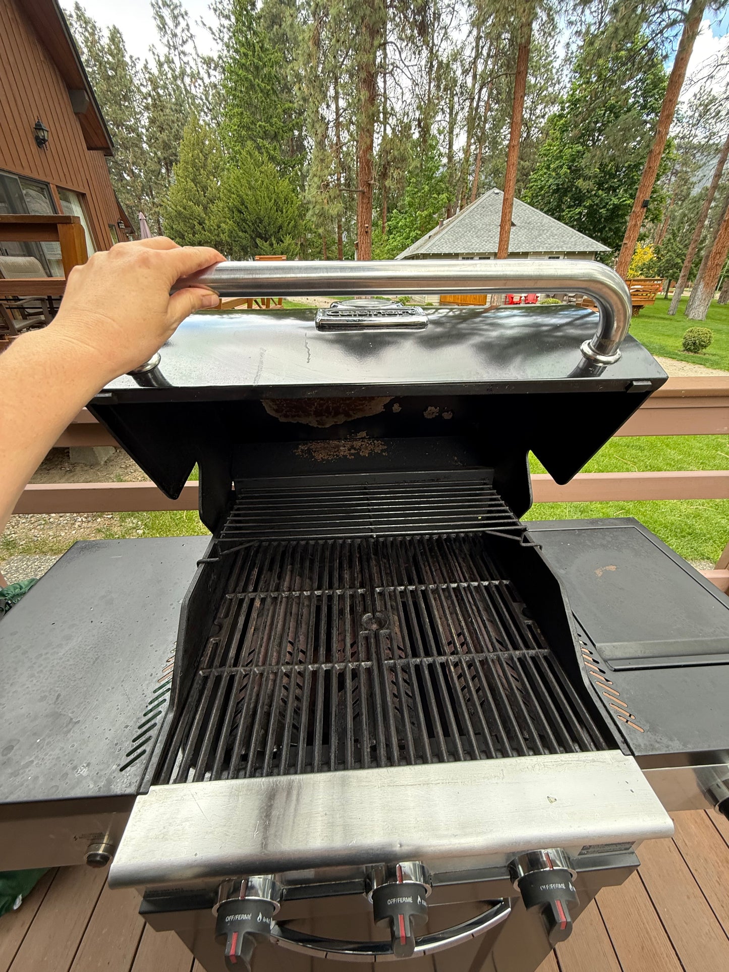 We see the hand of a professional cleaner holding open afreshly cleaned barbecu which is silver and gleaming, the knobs and the interior grate are all clean and fresh lookiing. In the background we can see trees and the side of a cabin.
