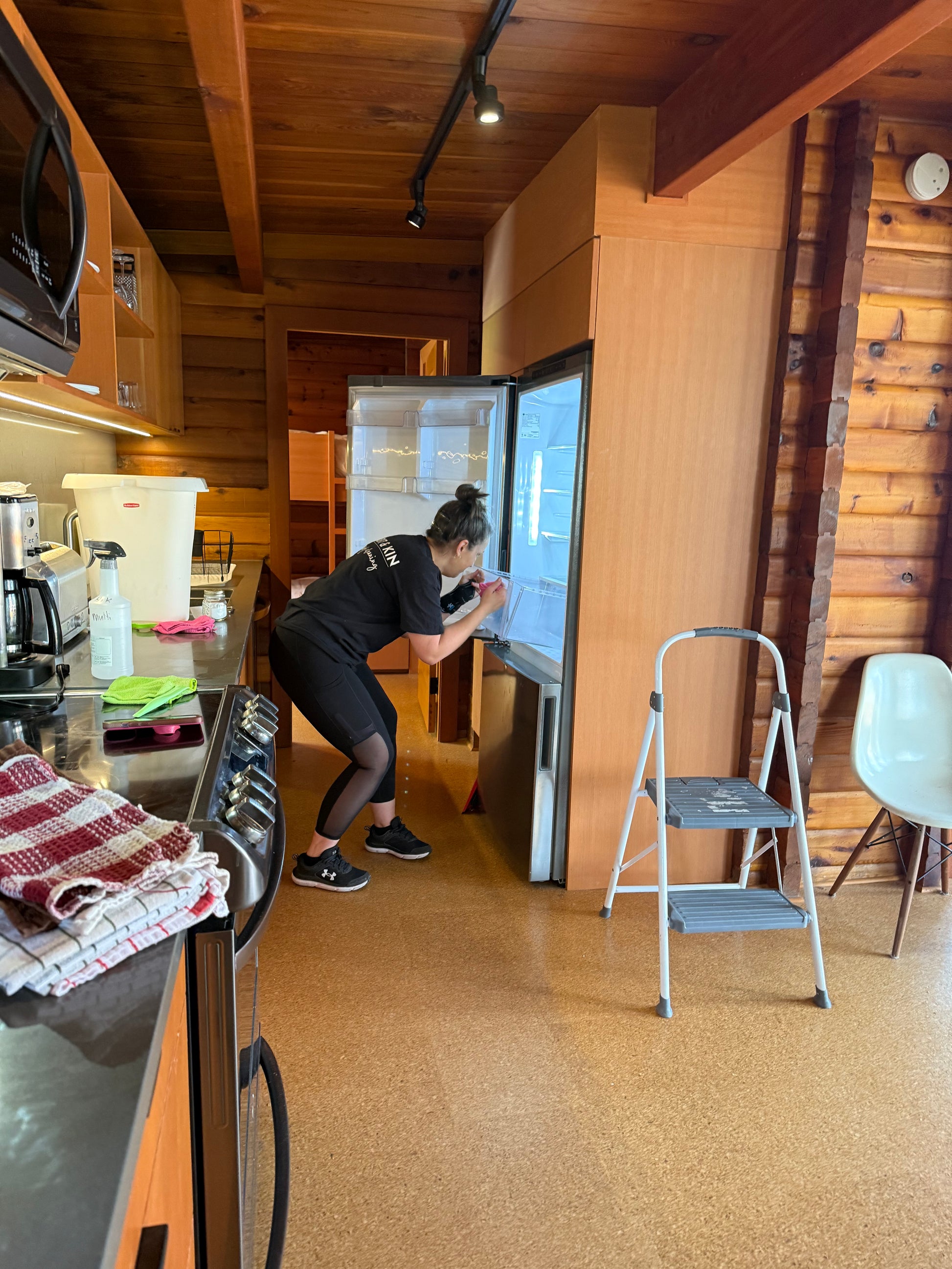 In the kitchen we see a professional cleaner from the side with an open fridge door replacing the crisper drawer after the inside has been cleaned to perfection. To the left we can see part of a counter with cleaning products on it, a stove and to the right a small stepp ladder in a room that looks like a cabin with the walls and ceiling being made of wood.