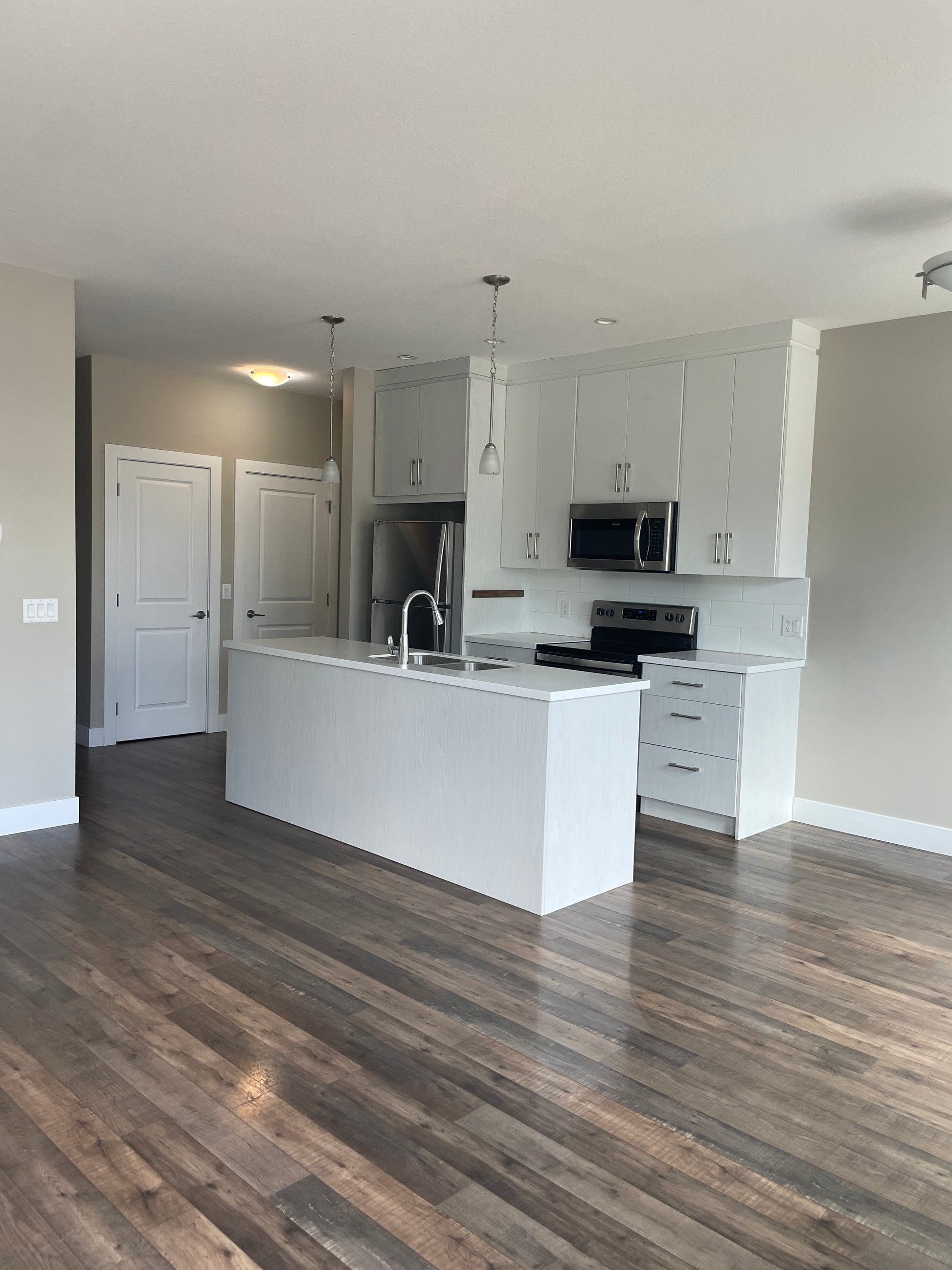 The picture appears to be taken from the living room with freshly cleaned laminate dark floors where there is no furniture and in the back we can see a kitchen with white counters and cupboards, a silver faucet, stainless steel oven, fridge and microwave and two doors.