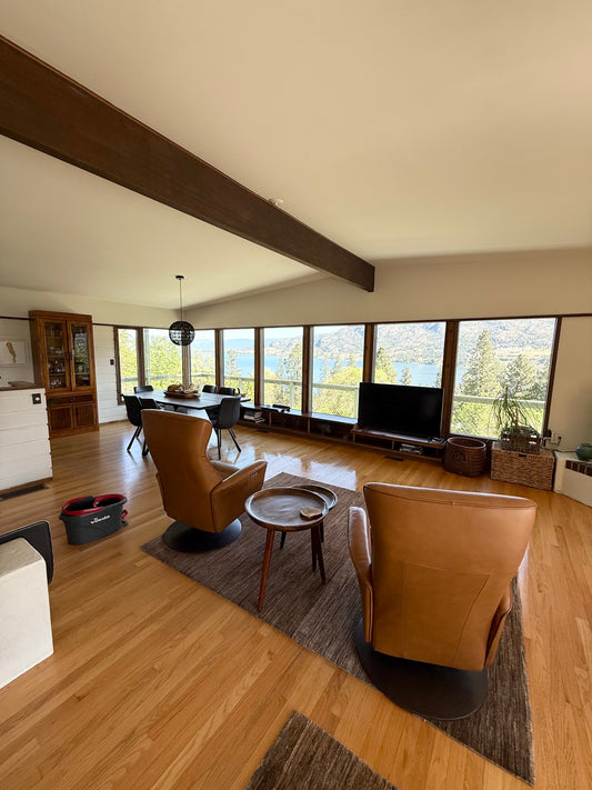 A picture taken of a living room with hardwood floors viewing a wall of windows and a beam in the ceiling with two brown leather chairs facing the windows and a table in between them.