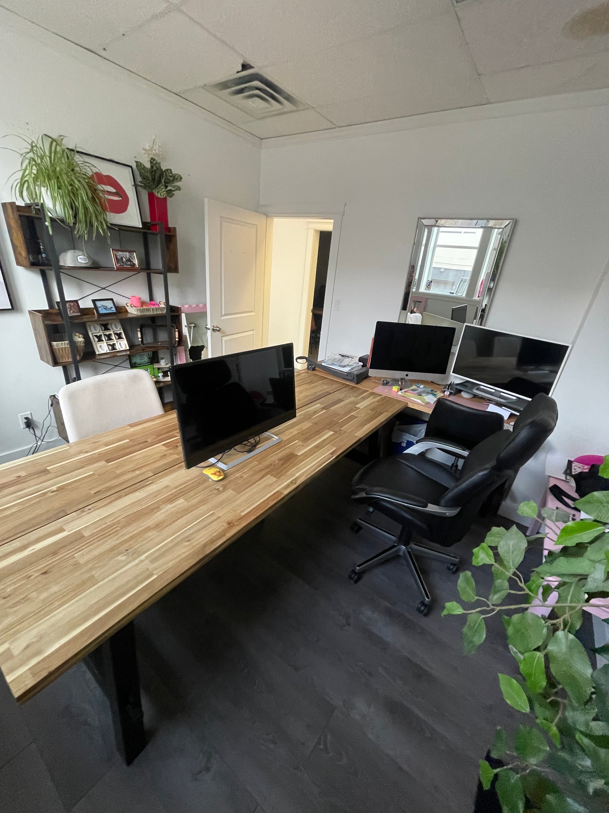 This picture is taken from behind the desk in an office with laminate flooring and a wodden desk that is freshly cleaned and cleared of debris with a computer monitor on it that has been cleaned of marks. To the right of the main desk is a smaller desk that has 2 monitors on it that are so clean you can see the reflection of a black rolling chair that is in front of them. The door can be seen in the distance as well as a shelf that has plants and pictures on it.