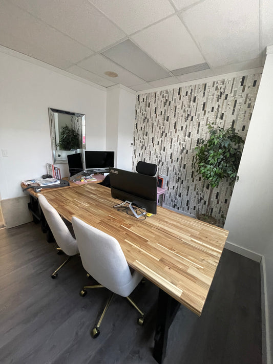 A picture of along desk in an office that has two chairs on one side for visitorsand a chair on the other side with a computer screen facing that chair and then another computer screen to the left of that and a patterned wallpaper on teh wall with a hanging plant and laminate floors all freshly cleaned.