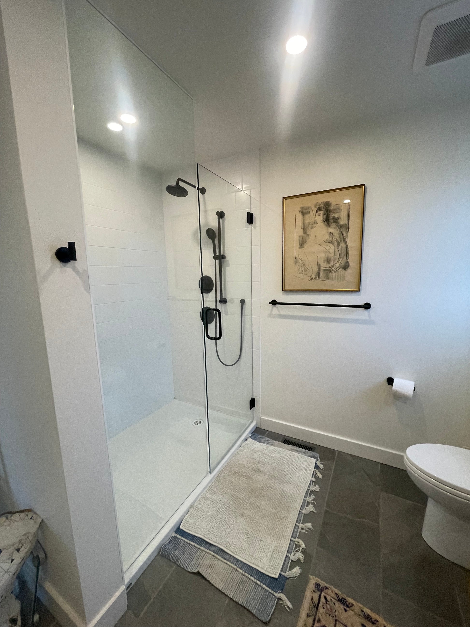 A picture of a stand up shower with white tiles and black fittings and a clear glass opening freshly cleaned with no marks or grim, dark tiled floor and a mat on the outside of the shower.