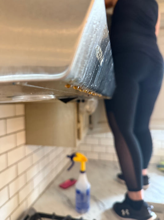 This phot is an up close shot of the oven hood which has old drips of grease on it, we catch a glimpse of the stovetop burners on the bottom and slightly blurry in the distance can see cleaning product spray, kitchen counters and a professional cleaner standing on the counter. 